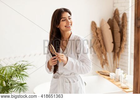 Brunette Woman Brushing Hair With Wooden Hairbrush In Modern Bathroom