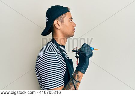 Young handsome african american man tattoo artist wearing professional uniform and gloves holding tattooer machine looking to side, relax profile pose with natural face and confident smile. 