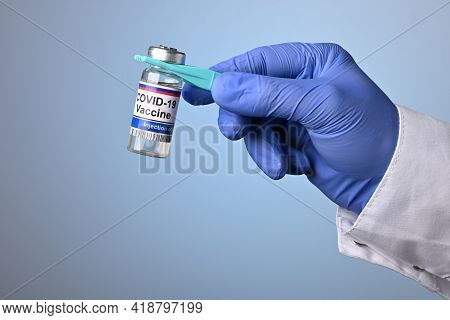 Doctor Hand Holds Covid-19 Vaccine Jar With Plastic Pincet. Hand In Blue Glove Offers Medical Treatm