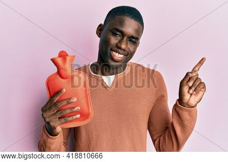 Young african american man holding heat water bag smiling happy pointing with hand and finger to the side 
