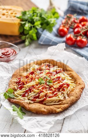 Tasty Deep-fried Langos Served On Baking Paper With A Lot Of Grated Cheese, Ketchup And Fresh Herbs