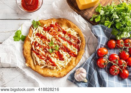 Tasty Deep-fried Langos Served On Baking Paper With A Lot Of Grated Cheese, Ketchup, Fresh Herbs And