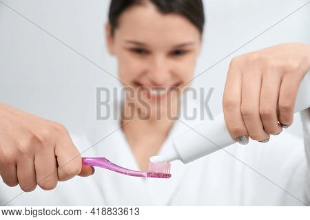 Front View Of Young Smiling Woman In White Robe Squeezes Toothpaste On Brush At Home. Concept Of Pre