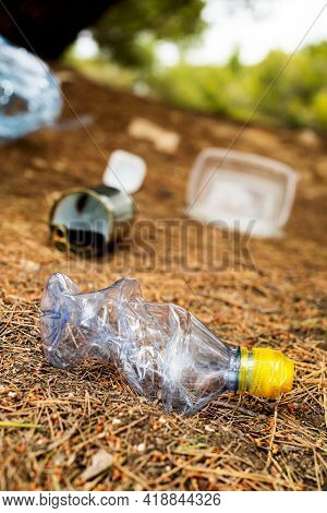 closeup of a used plastic bottle and some other garbage, such as used food cans and plastic containers, thrown on the ground of a forest