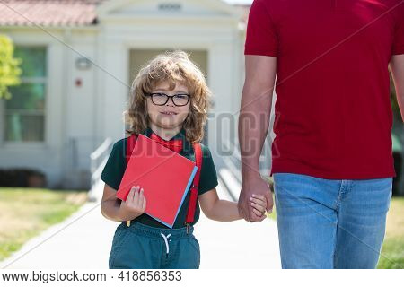 Parent And Nerd Pupil Of Primary School Go Hand In Hand. Father Holding Hand Of Son With Backpack Ou