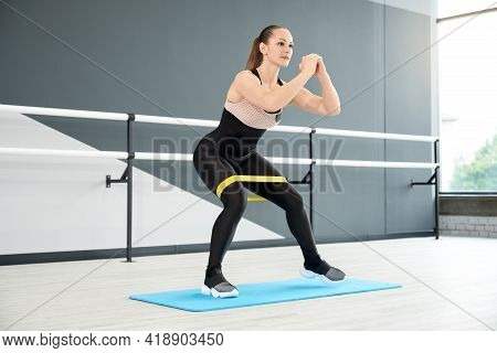 Side View Of Adult Woman In Tight Black Sportswear Using Fitness Band And Walking In Half Squat. Mus