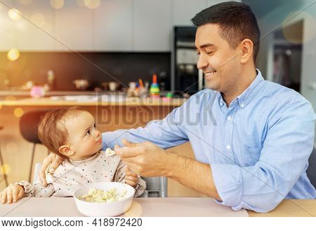 family, food, eating and people concept - happy smiling middle-aged father feeding little baby daughter sitting in highchair with puree by spoon at home
