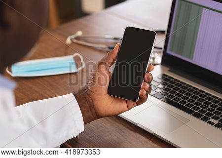 Mid section of african american senior male doctor holding smartphone with copy space at home. distant communication and telemedicine consultation concept.