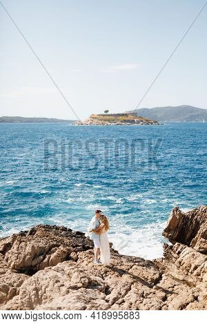 Man And Woman In Love Stand Embracing And Holding Hands On The Rocky Seashore, Behind Them Is The Ma