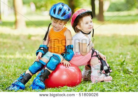 Little boy and girl in roller equipment sit together on red ball for jumping in summer park