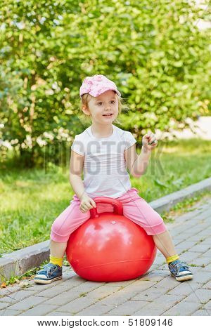 Smiling little girl sits on red ball for jumping on walkway in park, holding green leaf in her hand 