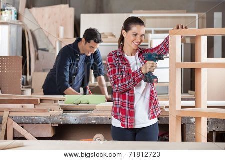 Happy female carpenter drilling wood while colleague working in background at workshop