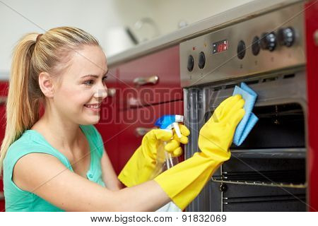 people, housework and housekeeping concept - happy woman with bottle of spray cleanser cleaning oven at home kitchen
