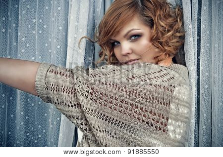 Mysterious trendy woman with short curly red hair standing between white long dotted lace curtains.