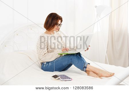 Young woman reads from laptop computer in bed. Engrossed Girl in eyeglasses and earphones with mobile phone. Attractive woman in casual clothes at white bed. Internet freelancer. High key, soft tone