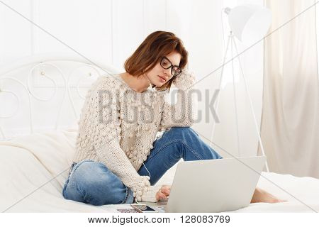 Young woman reads from laptop computer in bed. Engrossed Girl in eyeglasses and earphones with mobile phone. Attractive woman in casual clothes at white bed. Internet freelancer. High key, soft tone