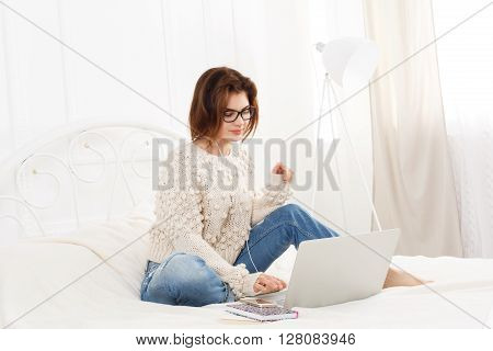 Young woman reads from laptop computer in bed. Engrossed Girl in eyeglasses and earphones with mobile phone. Attractive woman in casual clothes at white bed. Internet freelancer. High key, soft tone