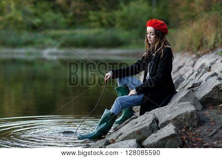 The girl in a red cap sits at a pond on stones. The girl holds a twig. The girl plays with water. The girl is dressed in green boots.