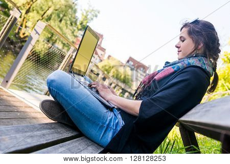 Young woman using laptop at a riverbank. Milleanial working in a park.