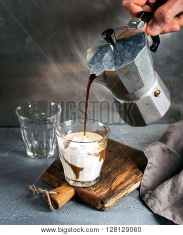 A glass of coffee with ice cream on a rustic wooden board. Drink is poured from a steel Italian Moka pot held by man's hand, grey background