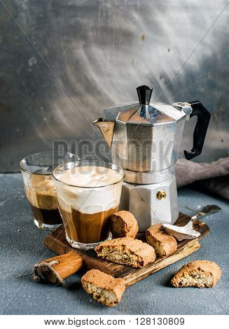 Glass of latte coffee on rustic wooden board, cantucci biscuits and steel Italian Moka pot, grey background, selective focus
