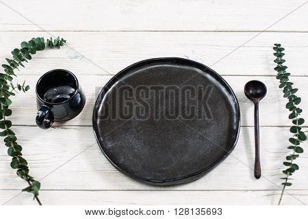 Top view empty black dishes and cutlery on rustic wooden table. Ceramic black plate, cup and spoon on white wooden background. Flat lay of handmade black ceramics on white wooden table.