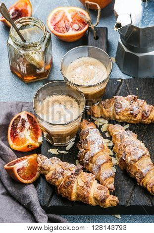 Italian style home breakfast. Latte coffee, almond croissants and red bloody oranges on dark wooden serving  board over concrete textured table, moka pot and honey jar at background, selective focus
