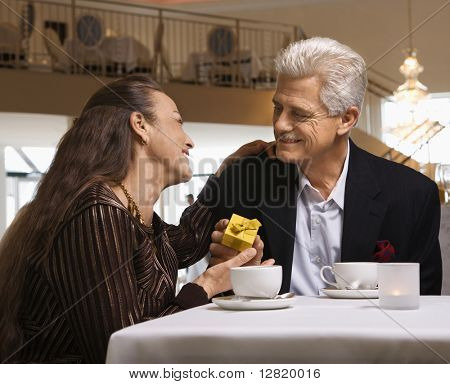Caucasian mature adult male giving prime adult female gift box at restaurant table.