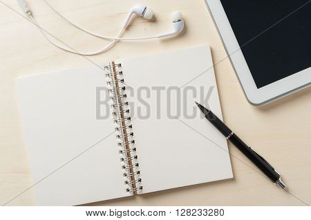 Notebook with blank area for text or message and pen beside small tablet pc and in ear headset on wood table
