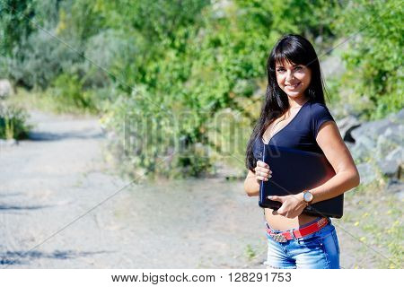 smiling brunette girl with long hair holding laptop standing on a mountaiin road. Looking to camera Sunny summer day