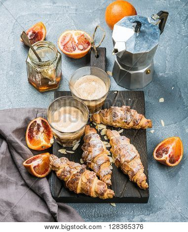 Italian style home breakfast. Latte coffee, almond croissants and red bloody oranges on dark wooden serving  board over concrete textured table, moka pot and honey jar at background, selective focus