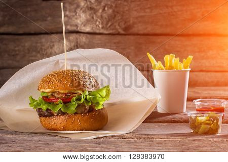 Burger on paper and fries. Table with beefburger and fries. Sliced pickles and fast food. High-calorie lunch at restaurant.