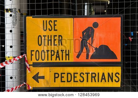 A sign for pedestrians to Use Other Footpath in yellow and orange color attached to metal net fence. 