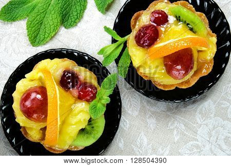 Cakes with slices of fresh fruit orange, kiwi and grapes with jelly and cream on a black saucers on a table cloth.