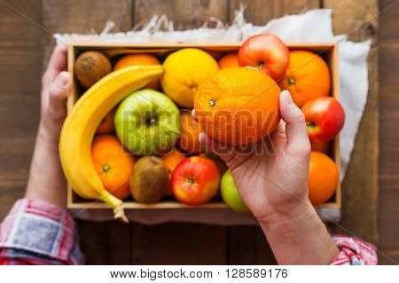 Man in tartan plaid shirt holds a box full of fresh fruits and a big juicy orange. Fruit harvest - apples oranges lemon kiwi banana. Rustic wooden table.