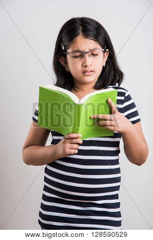indian small girl with eye glasses standing and reading a book, asian little girl holding a book and reading over white background, cute indian girl wearing spectacles and reading book while standing