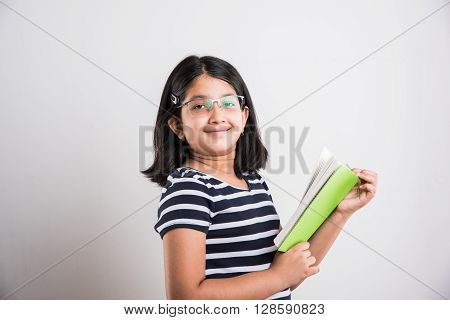 indian small girl with eye glasses standing and reading a book, asian little girl holding a book and reading over white background, cute indian girl wearing spectacles and reading book while standing