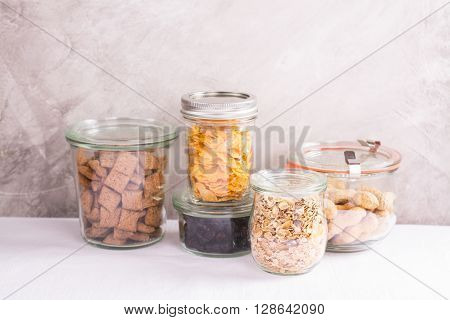 Assortment of cereals, flakes, nuts and berries in storage glass jars over kitchen table. Homemade granola bar ingredients