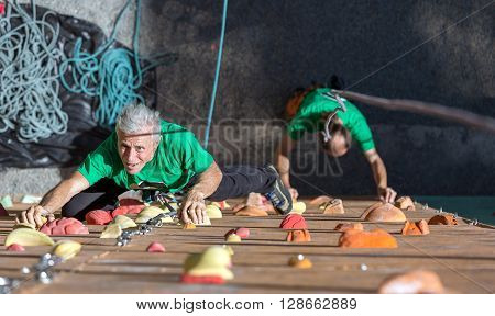Elderly Female Moving Up on Outdoor Climbing Wall Sporty Clothing on Fitness Training Intense but Positive Face another Climber Start His Ascent on Background