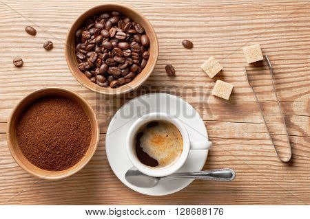 Coffee beans, ground coffee and cup of brewed coffee on rustic wooden table with sugar tongs, cane sugar cubes and coffee beans, view from above