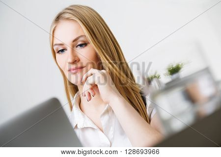 Portrait of attractive smiling caucasian businesswoman with hand at chin sitting at office desk with laptop and glass of water