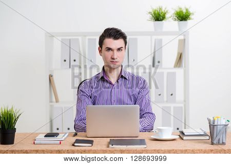 Caucasian businessman in striped purple shirt using laptop placed on office desktop and looking at the camera