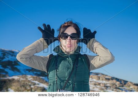 portrait of pretty face woman with green vest and sunglasses looking gesturing hands gloves making fun scoff and sticking out tongue in winter outdoor blue sky