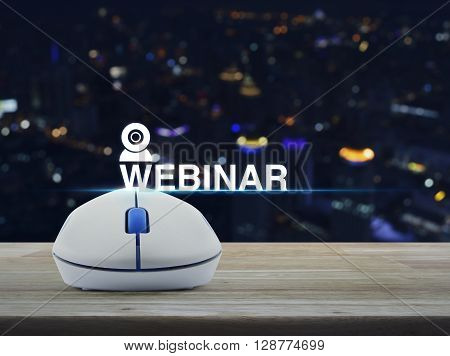 Wireless computer mouse with webinar icon on wooden table in front of blurred light city tower Seminar online concept