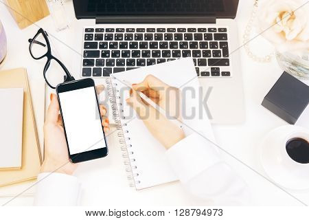Closeup of woman hands copying data from white mock up smart phone to notepad on designer desktop with laptop and coffee cup. Business accountant working on report via cell phone on desktop. Mock up