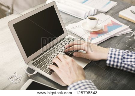 Man using two hands to type on laptop with blank screen on wooden desktop with business report and coffee cup. Mock up