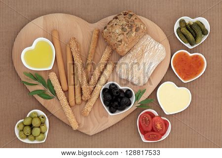 Crudities fresh snack selection with bread sticks and rolls, mayo and  sweet chilli dips, olives, oil, gherkins and tomatoes on a heart shaped wooden board