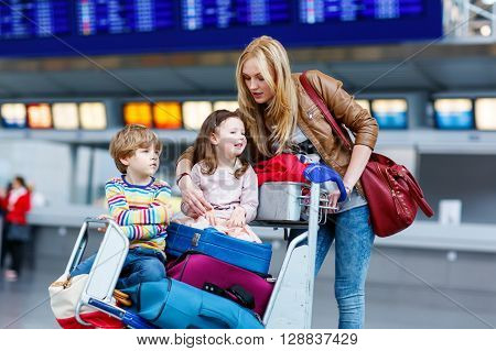 Two adorable little siblings, kid boy and girl sitting on suitcases on international airport. Young mother pushing. Brother and sister, happy family wating for flight and going on vacations.