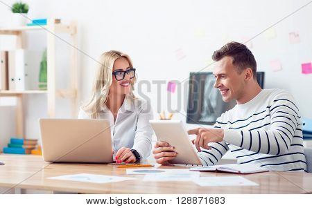 Dependant on modern technologies. Positive delighted smiling colleagues  sitting at the table and discussing project while being involved in work
