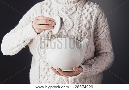 Woman In Warm Sweater Holds Big White Ceramic Teapot On One Hand And Opens Cap With Other. Front Vie